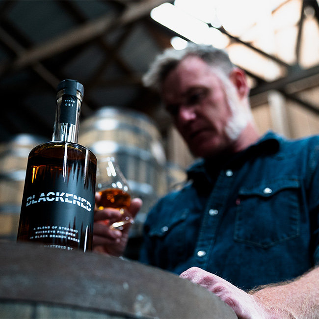 A Bottle of BLACKENED with Master Distiller Rob Dietrich looking into a Glencairn glass in the back