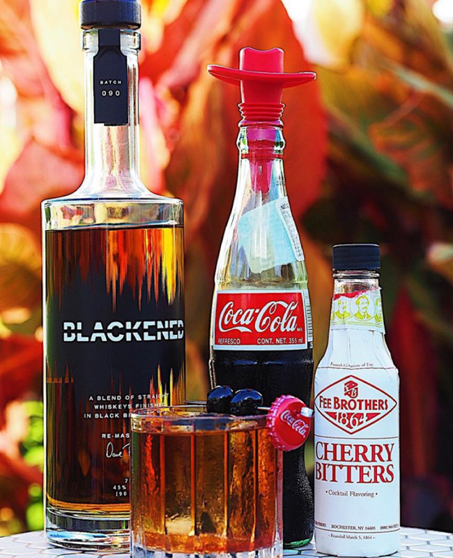 BLACKENED Coke Whiskey Cocktail in front of a bottle of blackened, cherry bitters, and Coke bottle