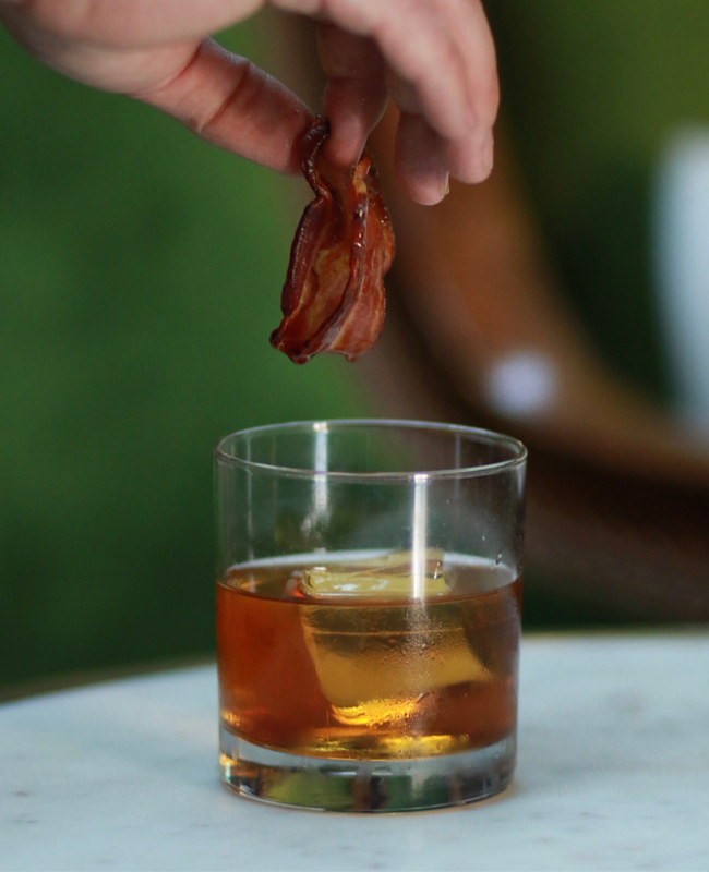 Hand holding bacon over BLACKENED's For Whom the Bell Tolls Whiskey Cocktail