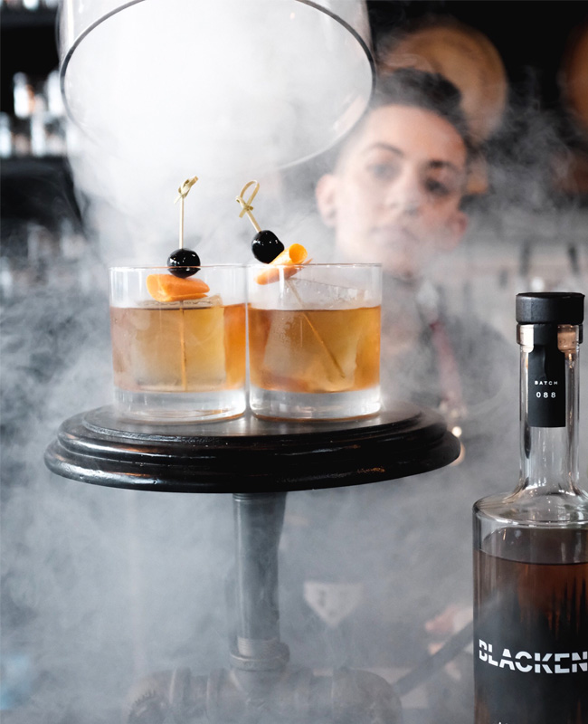 Bartender the pulling cover off two fresh Smoked Old Fashioned Cocktails