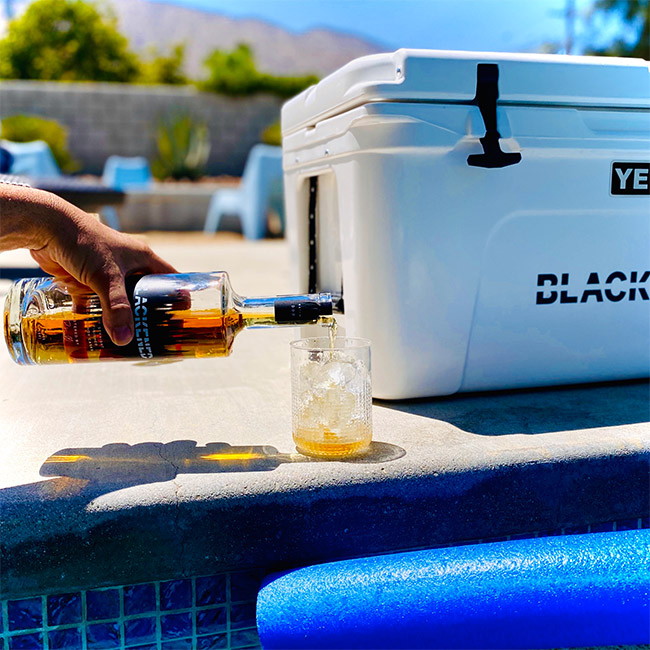 Person Pouring a Glass of BLACKENED Whiskey next to a pool in front of a YETI cooler